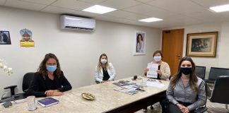 as quatro mulheres usando mascaras sentadas em volta de uma mesa olham para a câmera; adeliana mostra um papel