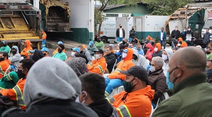 reunião de trabalhadores da comcap com uniformes, roupas de frio e máscaras, a maioria sentada, em pátio da empresa