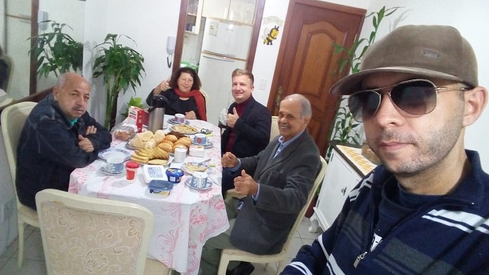 tiago faz selfie usando boné e óculos escuros e demais pessoas estão sentadas ao fundo em mesa com café posto em apartamento