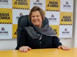 adeliana sorridente com as mãos sobre uma mesa; ao fundo painel com logos da prefeitura de são josé