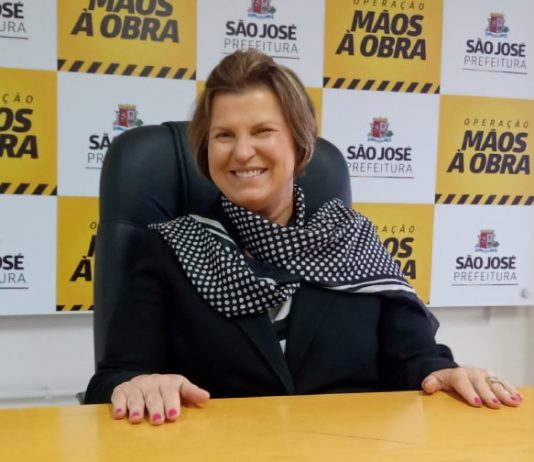 adeliana sorridente com as mãos sobre uma mesa; ao fundo painel com logos da prefeitura de são josé