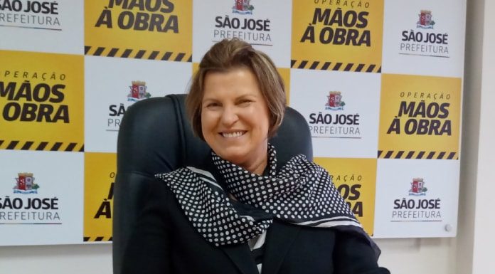 adeliana sorridente com as mãos sobre uma mesa; ao fundo painel com logos da prefeitura de são josé