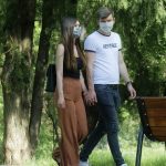 homem e mulher brancos, por volta de 30 anos, andam de máscaras e mãos dadas por parque, com banco e árvores ao fundo