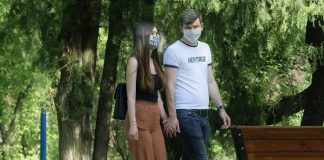 homem e mulher brancos, por volta de 30 anos, andam de máscaras e mãos dadas por parque, com banco e árvores ao fundo
