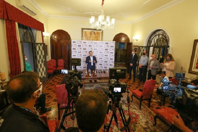 Homem de pé em uma sala dando entrevista para vários jornalistas