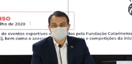 Novo pedido de impeachment contra Moisés é aberto na Alesc envolvendo compra de respiradores governador moisés usa máscara falando em um microfone, sentado atrás de mesa e ao fundo painel com informações