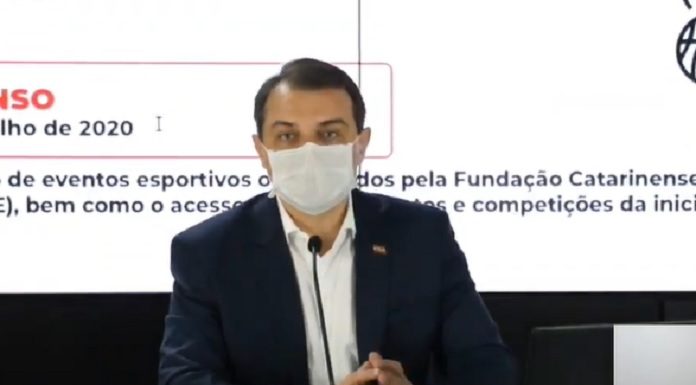 governador moisés usa máscara falando em um microfone, sentado atrás de mesa e ao fundo painel com informações
