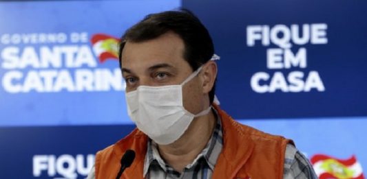 Suposta participação de Moisés na compra dos respiradores faz processo ir ao STJ carlos moisés usando máscara e colete da defesa civil em frente a um microfone e ao fundo painel do governo de santa catarina com inscrição "fique em casa"