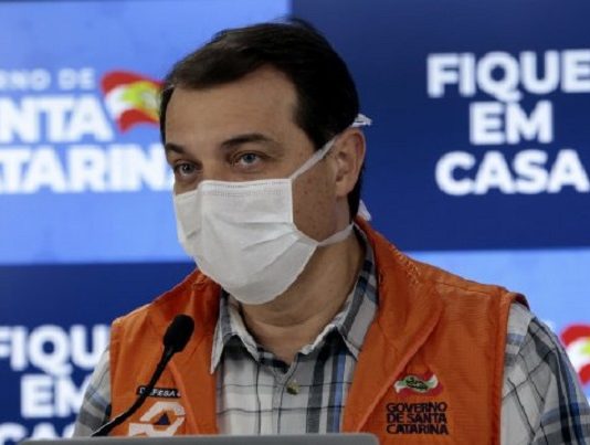 carlos moisés usando máscara e colete da defesa civil em frente a um microfone e ao fundo painel do governo de santa catarina com inscrição "fique em casa"