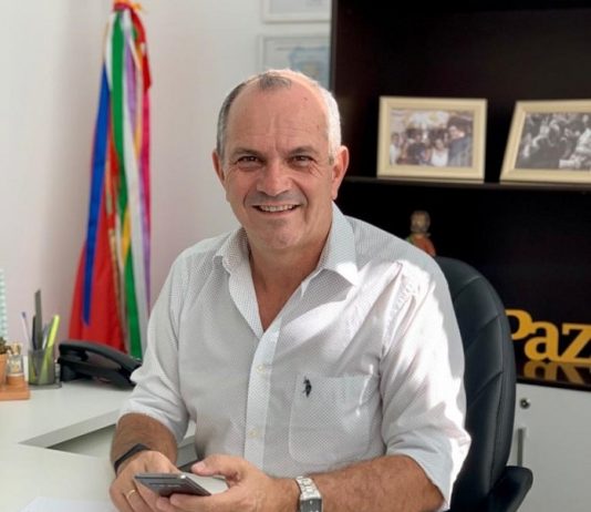 PDT de São José lança pré-candidatura à prefeitura moacir da silva sentado em seu gabinete olha sorridente para a câmera segurando um celular; fotos e bandeiras ao fundo