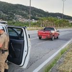policial mexendo dentro da viatura com porta aberta estacionada atrás do carro gm montana parado no meio da pista direita