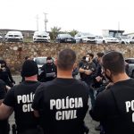 diversos policiais civis vistos de costas reunidos em pé em estacionado; nome da pc nas costas