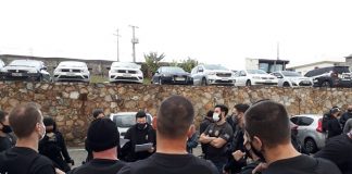 diversos policiais civis vistos de costas reunidos em pé em estacionado; nome da pc nas costas