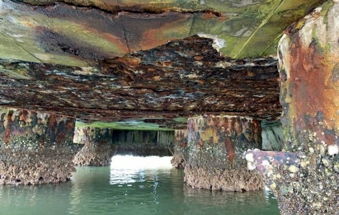 imagem da parte inferior de um bloco de pilares da pontes com concreto bastante deteriorado e cheio de cracas marítimas, pouco acima da superfície da água