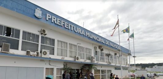 fachada da sede da prefeitura municipal de palhoça com pessoas andando na frente, motos estacionadas e três bandeiras