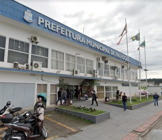 fachada da sede da prefeitura municipal de palhoça com pessoas andando na frente, motos estacionadas e três bandeiras