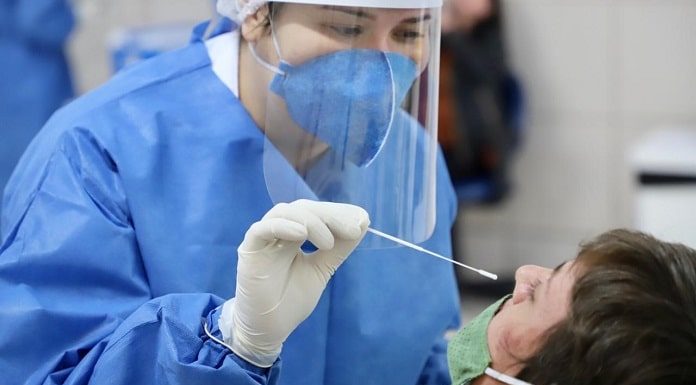 mulher vestindo jaleco azul, com luvas brancas fazendo o teste em homem usando máscara verde