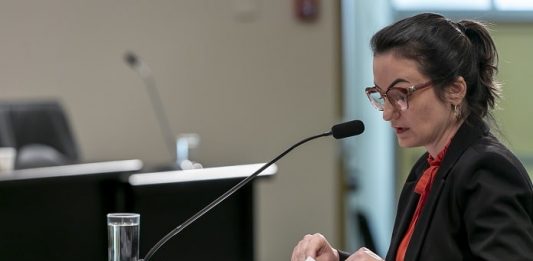 servidora sentada em mesa com microfone e papeis sobre a mesa