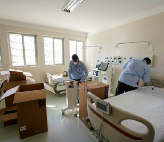 dois homens desencaixotam equipamentos dentro de quarto de enfermaria com duas camas
