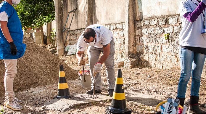 homem se abaixa para fazer inspeção em tampa de esgoto com cones em volta no meio de uma área em construção