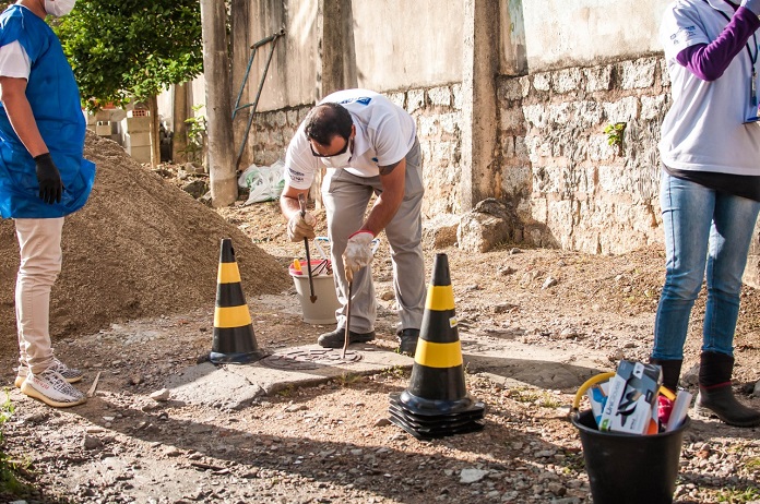 homem se abaixa para fazer inspeção em tampa de esgoto com cones em volta no meio de uma área em construção