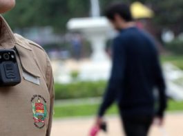 Justiça de SC determina que câmeras policias gravem automaticamente ocorrências close em câmera no uniforme de um policial em uma praça e um homem passando ao fundo