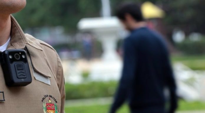 close em câmera no uniforme de um policial em uma praça e um homem passando ao fundo