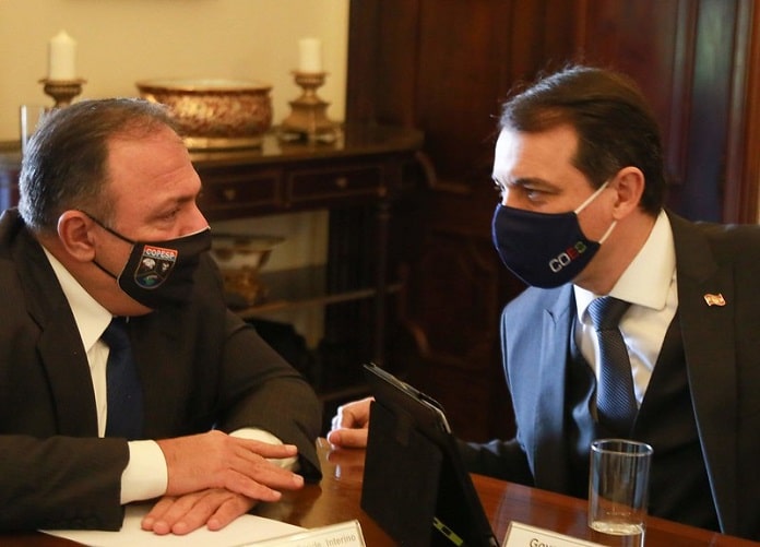 pazuello e moisés, ambos usando máscaras, conversam sentados na ponta de uma mesa