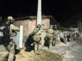 policias com uniformes camuflados e armamento pesado andando em calçada em frente a residências pobres em foto noturna
