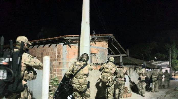 policias com uniformes camuflados e armamento pesado andando em calçada em frente a residências pobres em foto noturna