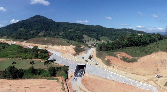 foto aérea de pequeno túnel em construção com morros ao fundo e caminhões por perto