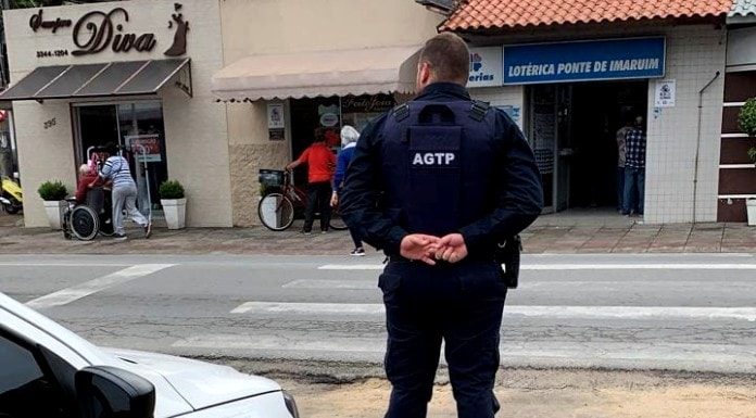 guarda de trânsito visto de costas com as mãos cruzadas para trás, olhando para rua, com carro estacionado ao lado