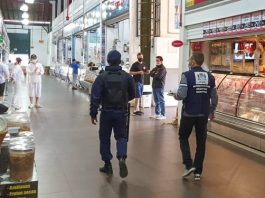 dois agentes da prefeitura andando no corredor do mercado público entre as lojas e pessoas em volta com máscaras