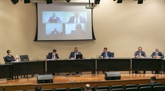 deputados membros da cpi sentados ao longo de bancada sobre palco de auditório com telão acima