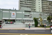 fachada do prédio do hospital vista do outro lado da rua; mulher passa na calçada