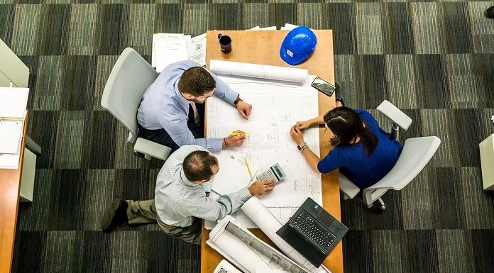 reunião de tres pessoas (dois homens e uma mulher) vistos de cima; estão em volta de uma mesa de escritório com notebook e papeis