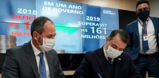 moisés e schuster usando máscara sentados lado a lado; moisés assina um papel sobre a mesa; telão ao fundo