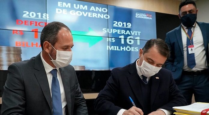 moisés e schuster usando máscara sentados lado a lado; moisés assina um papel sobre a mesa; telão ao fundo