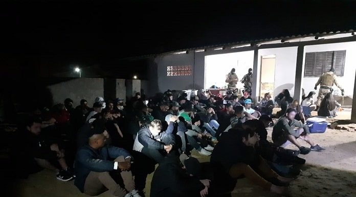 dezenas de homens sentados em pátio de casa com policiais em volta em foto noturna; alguns são iluminados pelas lanternas