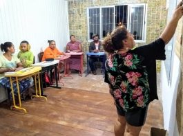 professora vista de costas dando aula apontando para o quadro em pequena sala com outras mulheres; há um garoto jovem com uma delas