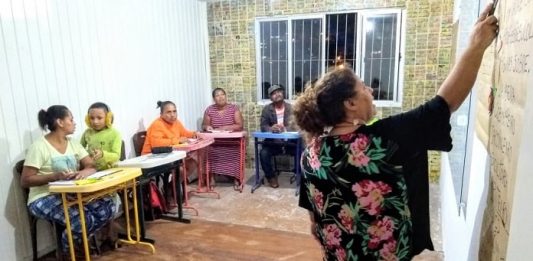 professora vista de costas dando aula apontando para o quadro em pequena sala com outras mulheres; há um garoto jovem com uma delas