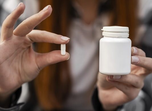 mulher segura pílula com dois dedos em uma mão e na outra o pote de remédio