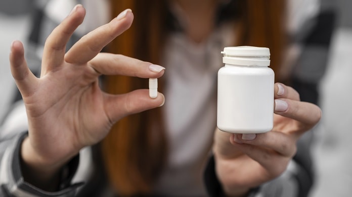 mulher segura pílula com dois dedos em uma mão e na outra o pote de remédio