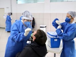 PMF se preocupa com exigência de testes da Covid-19 para retornar à Argentina duas mulheres de roupa azul, com toca na cabeça, usando máscara, fazendo teste em um homem de casaco preto, sentado em uma cadeira