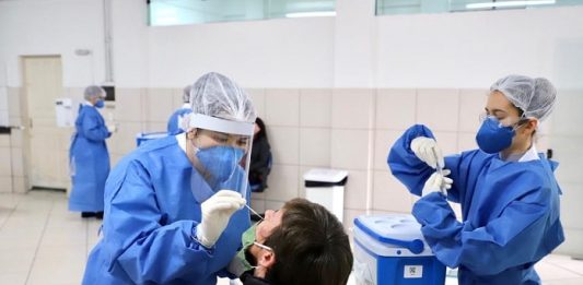 duas mulheres de roupa azul, com toca na cabeça, usando máscara, fazendo teste em um homem de casaco preto, sentado em uma cadeira