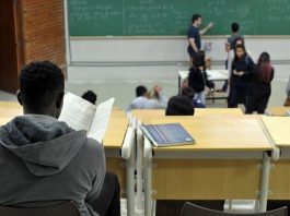 homem visto de costas em bancada de sala de aula com pessoas na frente da sala de aula, em pé em frente ao quadro negro