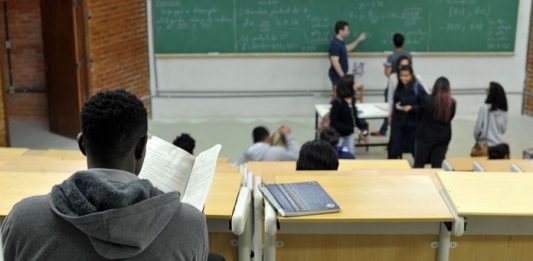 homem visto de costas em bancada de sala de aula com pessoas na frente da sala de aula, em pé em frente ao quadro negro