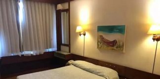 cama de casal em quarto de hotel arrumado com quadro de baleeira sobre a parede, cortina fechada e luzes acesas