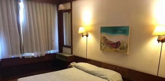 cama de casal em quarto de hotel arrumado com quadro de baleeira sobre a parede, cortina fechada e luzes acesas