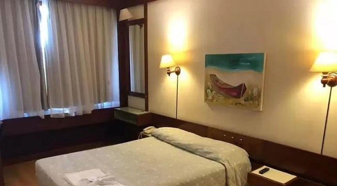 cama de casal em quarto de hotel arrumado com quadro de baleeira sobre a parede, cortina fechada e luzes acesas